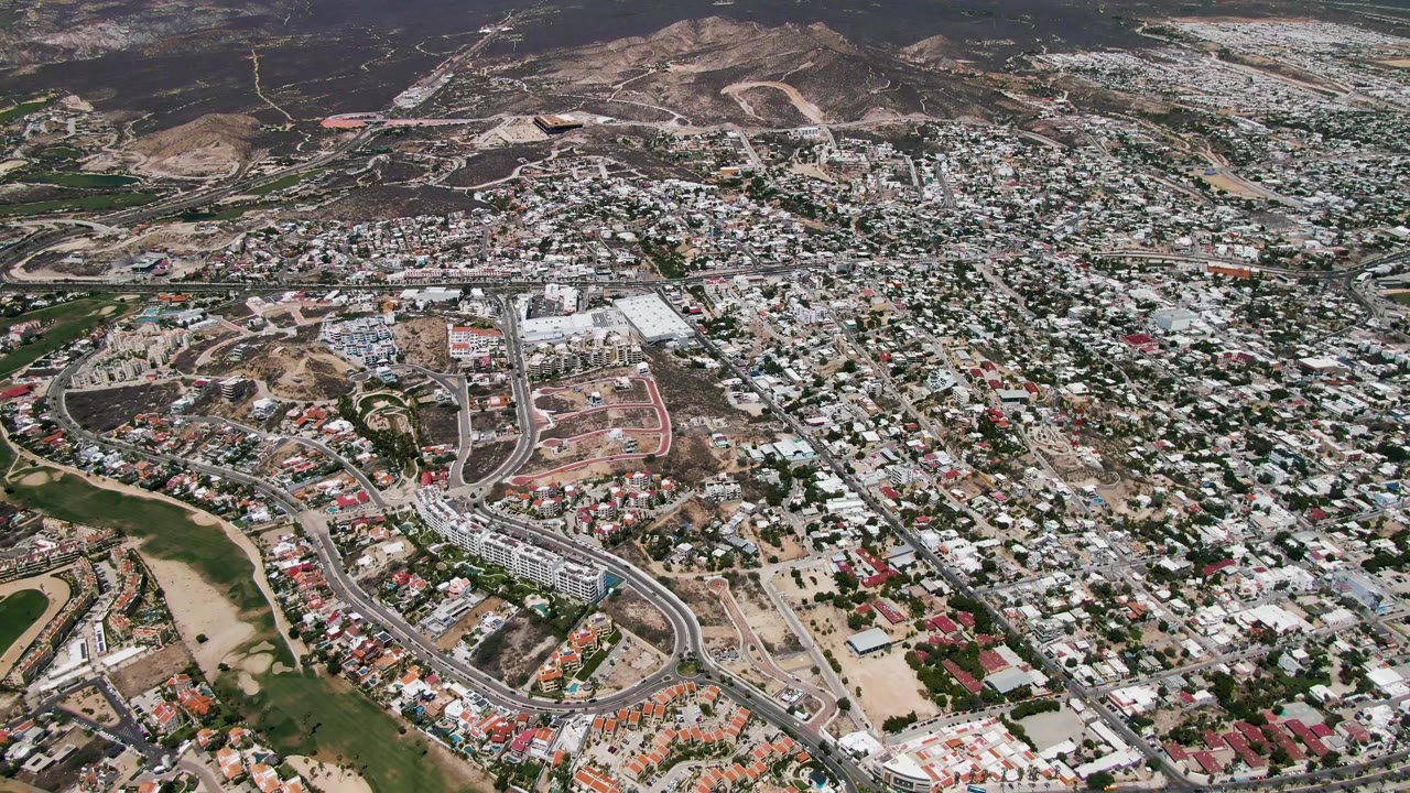 Mid-clip aerial view in San Jose Del Cabo showing a broad residential grid, larger roads, open tan land, green spaces, and hillside terrain, with no visible people.