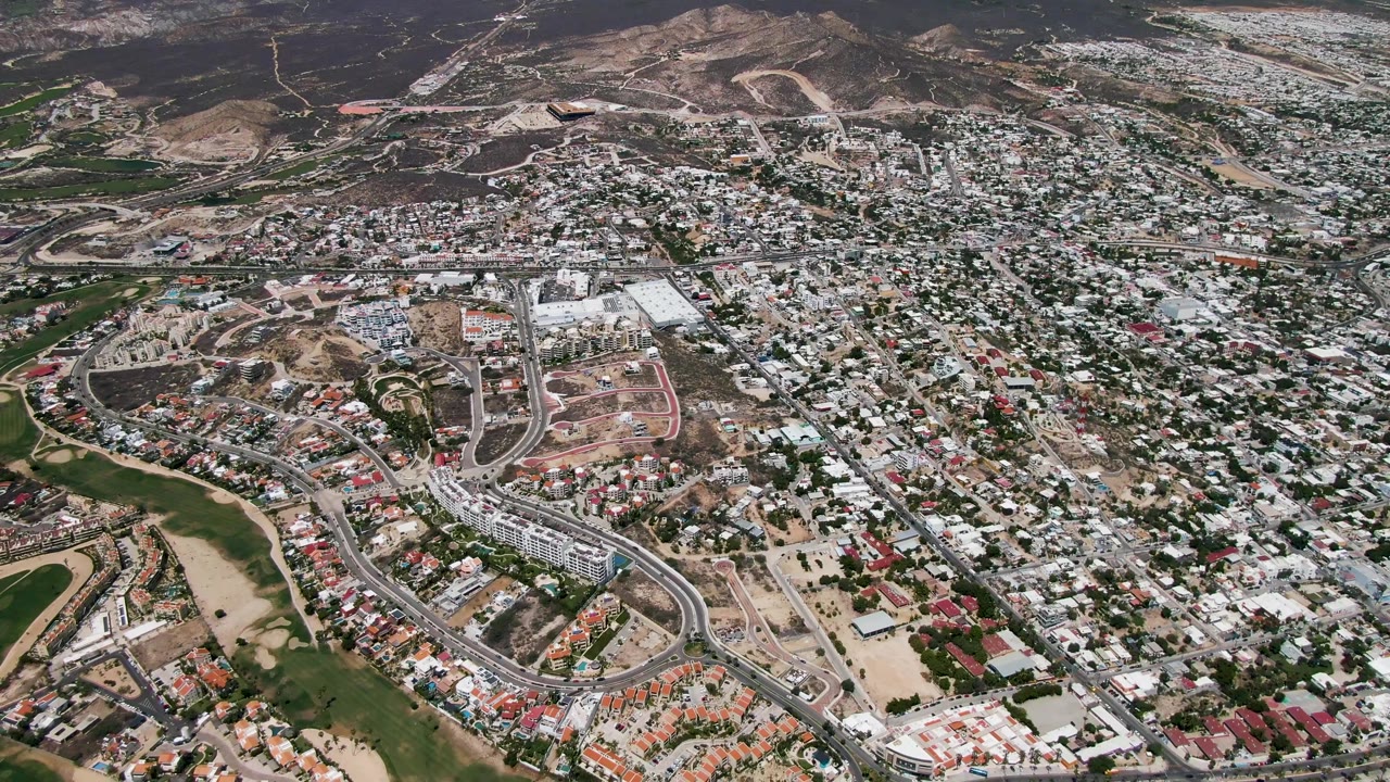 Aerial neighborhood frame with houses, road loops, green corridor, and dry hills.