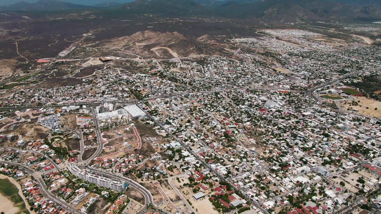 High aerial frame over San Jose Del Cabo with dense white-roofed residential blocks, curved roads, open brown lots, dry hills on the horizon, and no visible people.