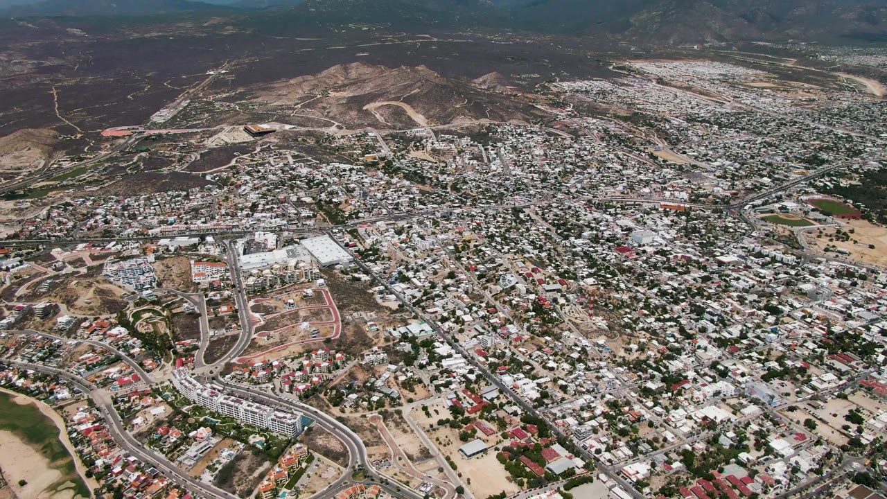 San Jose Del Cabo appears from a high drone angle with a broad residential grid, a looping road corridor near the lower frame, dry open land, and no visible people.