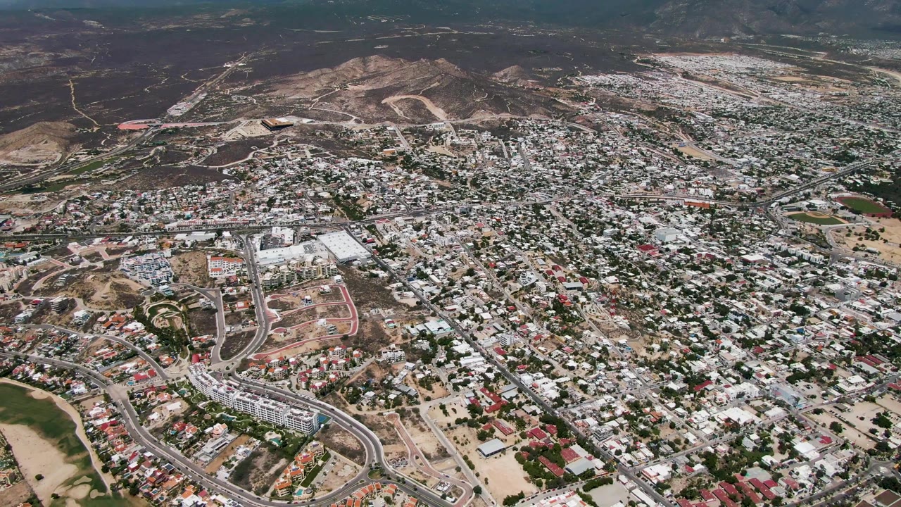 High aerial view of San Jose Del Cabo showing clustered homes, new-looking hillside road cuts, pale roofs, scattered red roofs, brown terrain, and no visible people.