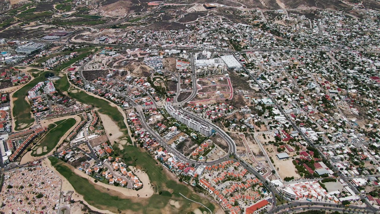 High aerial view over San Jose Del Cabo showing a green golf course, dense residential blocks, roads, dry lots, and no visible people.