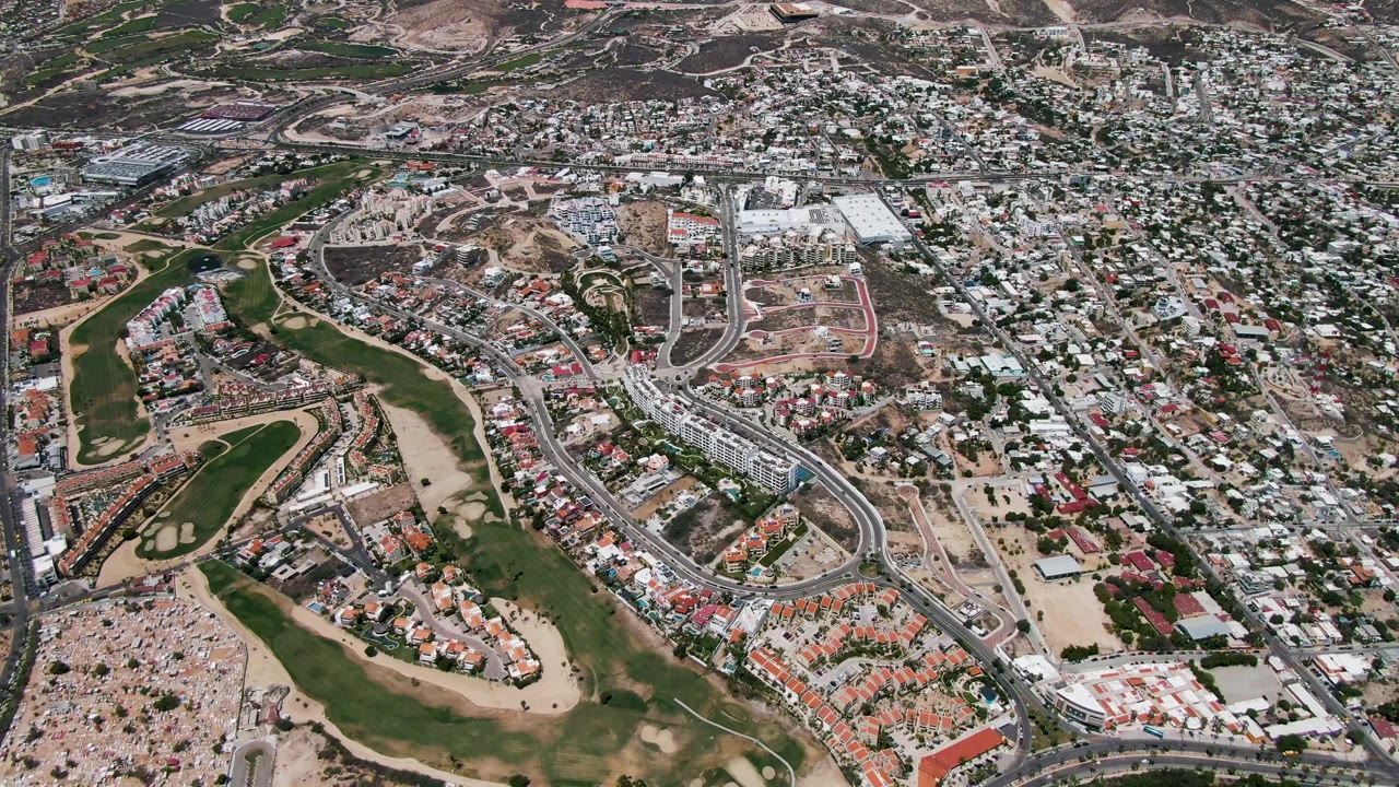 Aerial frame over San Jose Del Cabo with curving roads, red-roofed homes near the golf course, dense city blocks, construction areas, and no visible people.