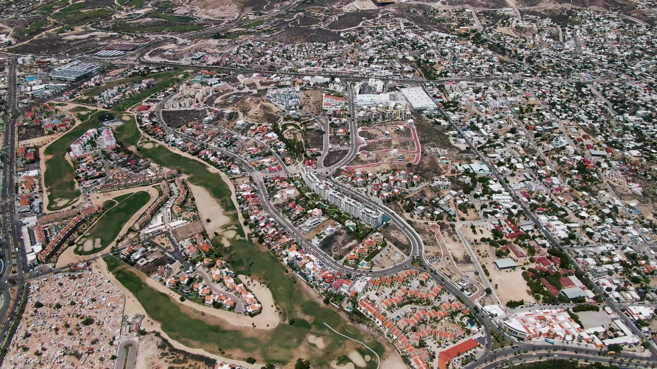 Wide drone perspective over San Jose Del Cabo showing neighborhoods packed around a golf course, paved roads, sandy open land, and no visible people.