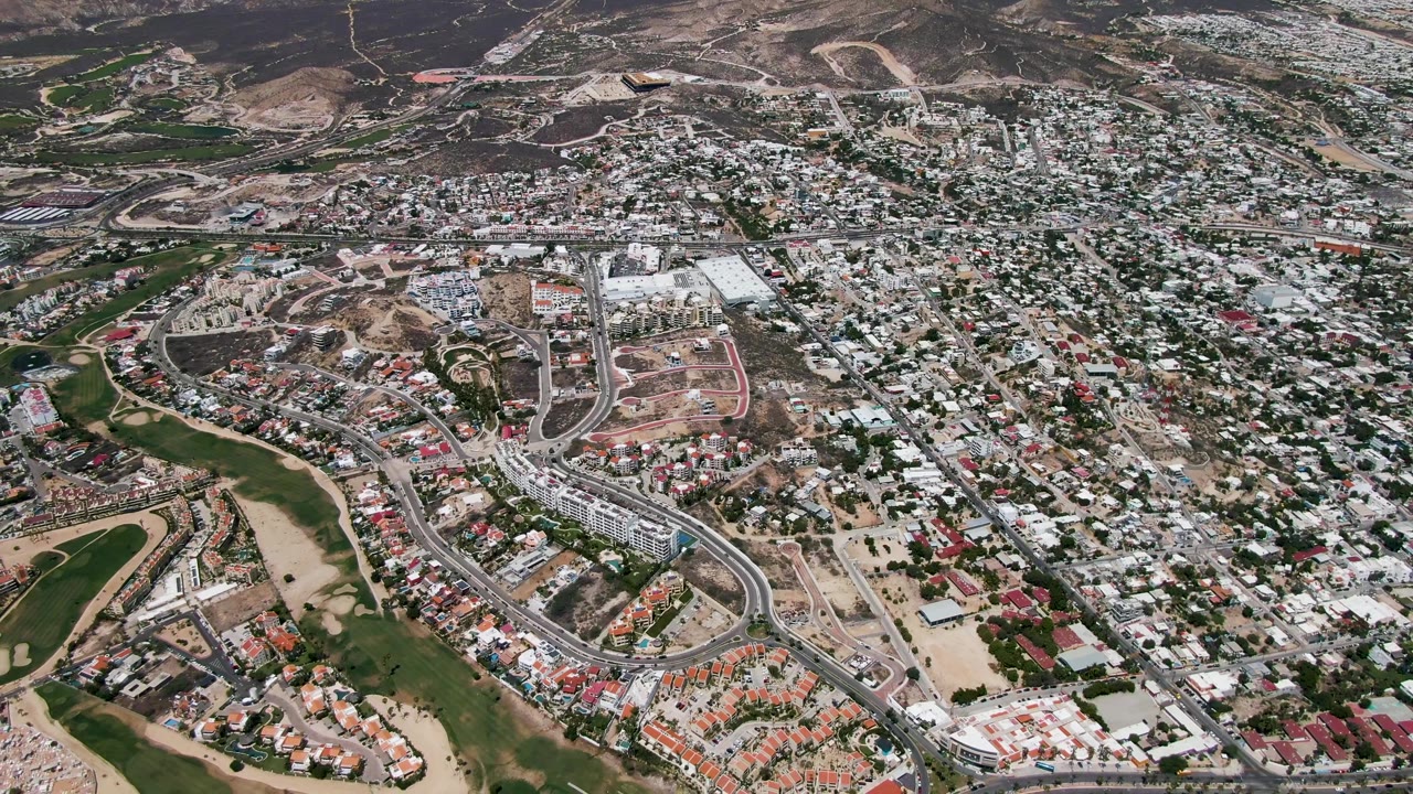 Aerial frame over San Jose del Cabo showing dense homes, curving roads, dirt construction areas, green open space, distant hills, and no visible people.