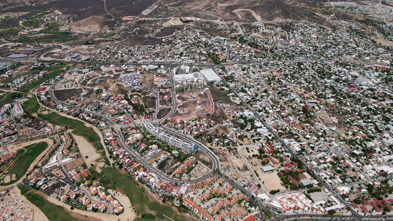 Aerial frame over San Jose del Cabo with a broad view of housing blocks, raw dirt development sites, paved roads, green areas, hills, and no visible people.