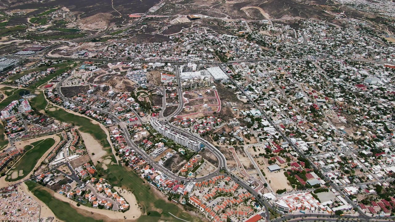 Aerial frame over San Jose del Cabo showing the city edge, residential streets, construction parcels, green open space, hills, and no visible people.