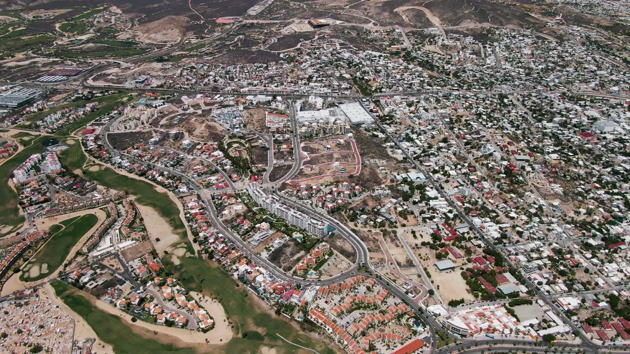 Oblique drone view of neighborhoods, green open areas, roads, dirt lots, and hills.