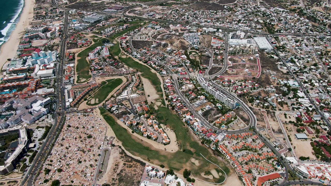 Aerial frame over San Jose Del Cabo showing surf along the left edge, a sandy beach, green golf fairways, roads, parking areas, and inland residential blocks with no visible people.