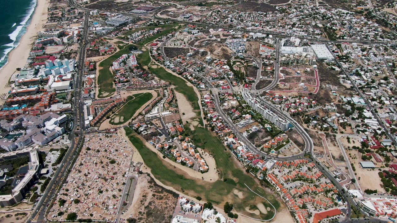 Aerial view in San Jose Del Cabo with ocean surf beside the beach, a curving golf course through the middle, multi-lane roads, parking lots, and residential buildings with no visible people.
