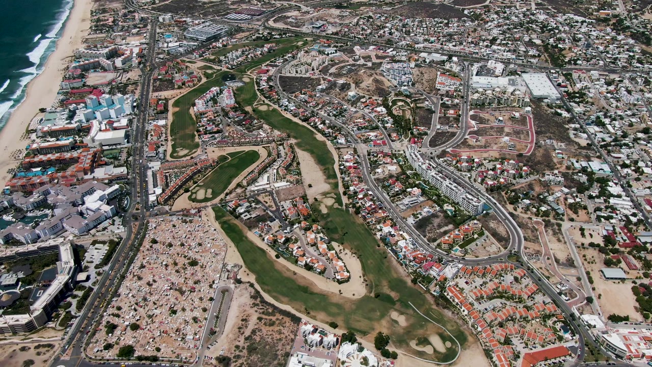 San Jose Del Cabo aerial frame showing the ocean and breaking waves near the beach, a green fairway corridor, a large parking area, roads, and dense housing with no visible people.