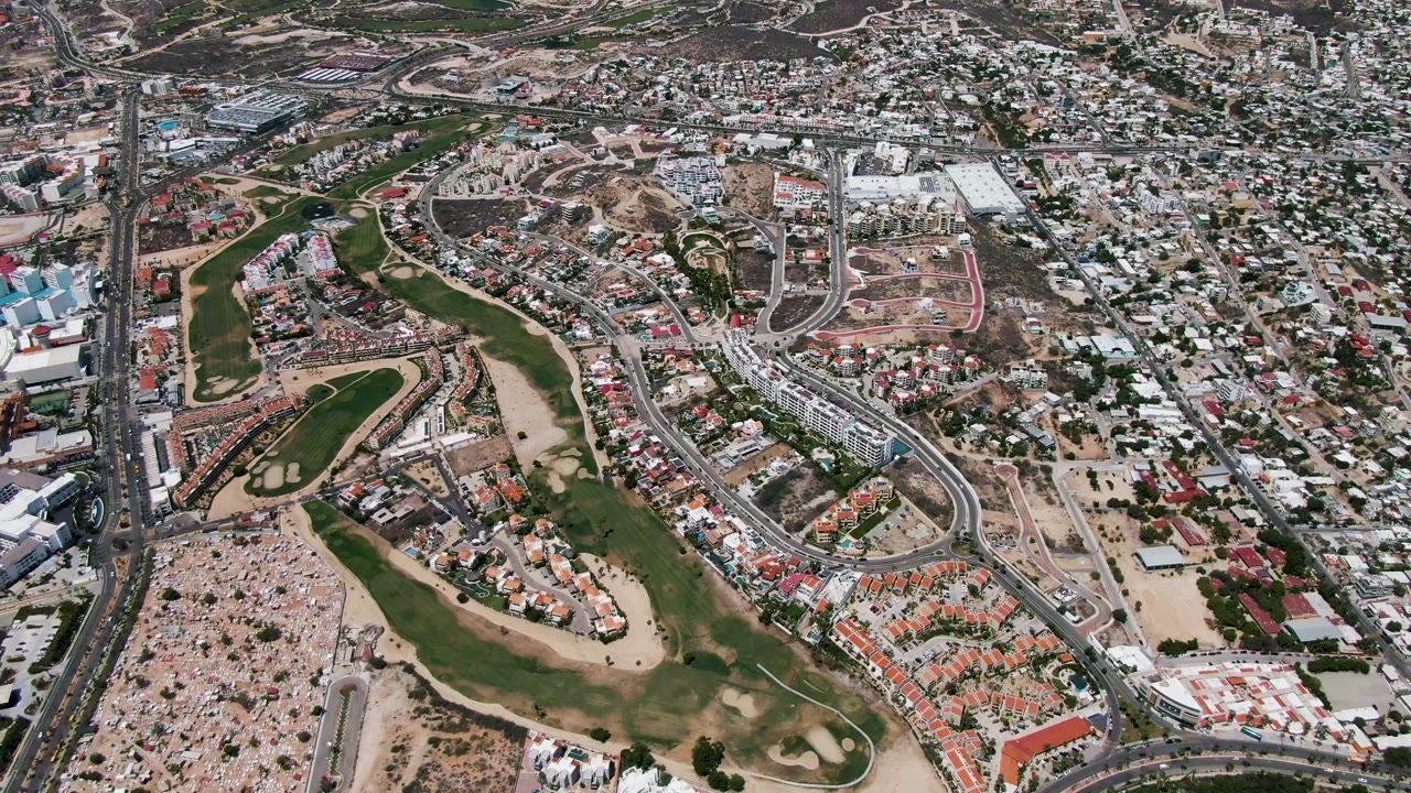 High aerial view over San Jose Del Cabo with curving roads, dense buildings, green golf course fairways, sandy ground, a strip of beach, blue ocean at the left edge, and no visible people.