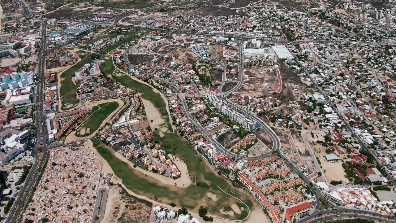 Oblique drone view of San Jose Del Cabo with the golf course centered, road corridors running through the scene, compact urban blocks to the right, beach and ocean along the left side, and no visible people.