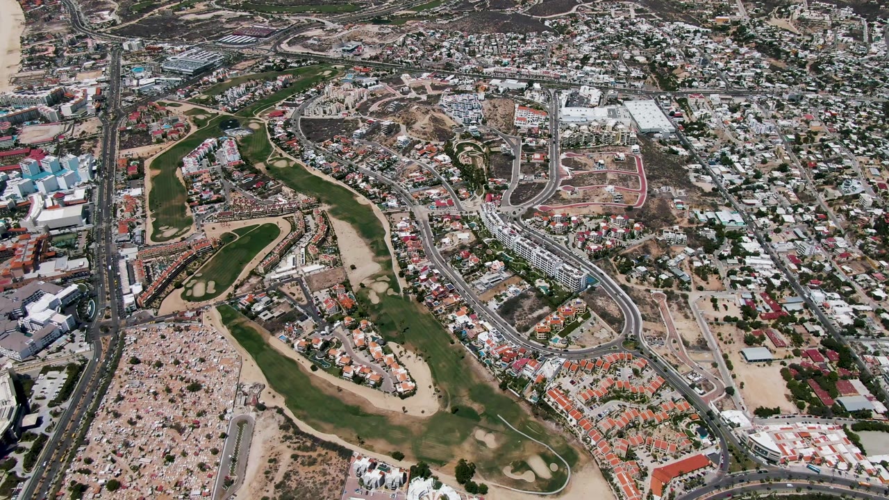 San Jose Del Cabo aerial frame showing fairways and sand traps in the lower half, a main road and dense buildings through the center, ocean water on the left, and no visible people.
