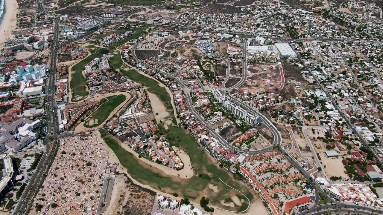Aerial city and coastline view with beach on the left, golf course below, and roads through buildings.