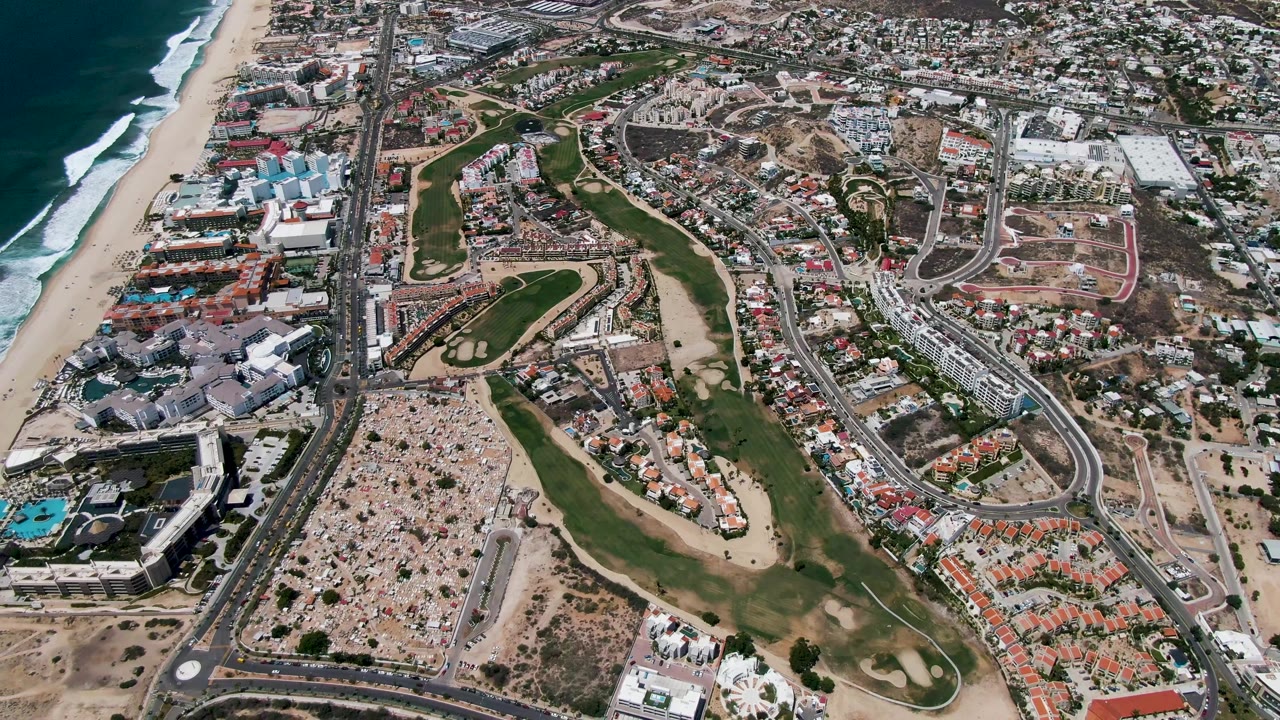 Aerial frame over San Jose Del Cabo shows surf on the left, a sandy beach, a coastal highway, green golf fairways, and dense buildings inland with no visible people.