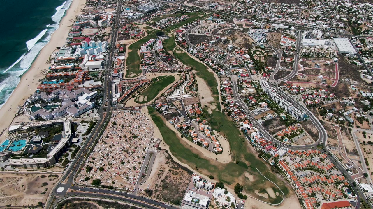 Aerial view in San Jose Del Cabo shows white surf beside the beach, large beachfront buildings, a curving road with vehicles, and a golf course running through the center with no visible people.
