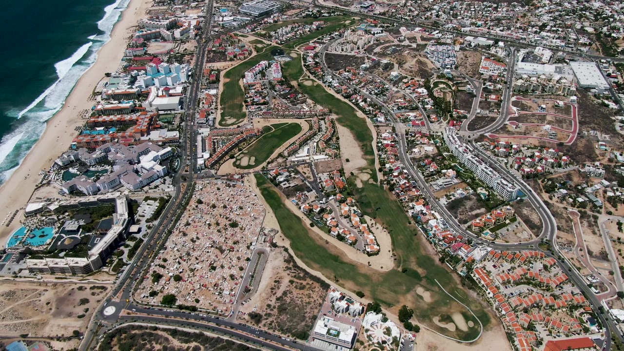 High aerial frame over San Jose Del Cabo places the beach and ocean along the left edge, with traffic on the highway, fairways and sand traps in the middle, and neighborhoods beyond with no visible people.