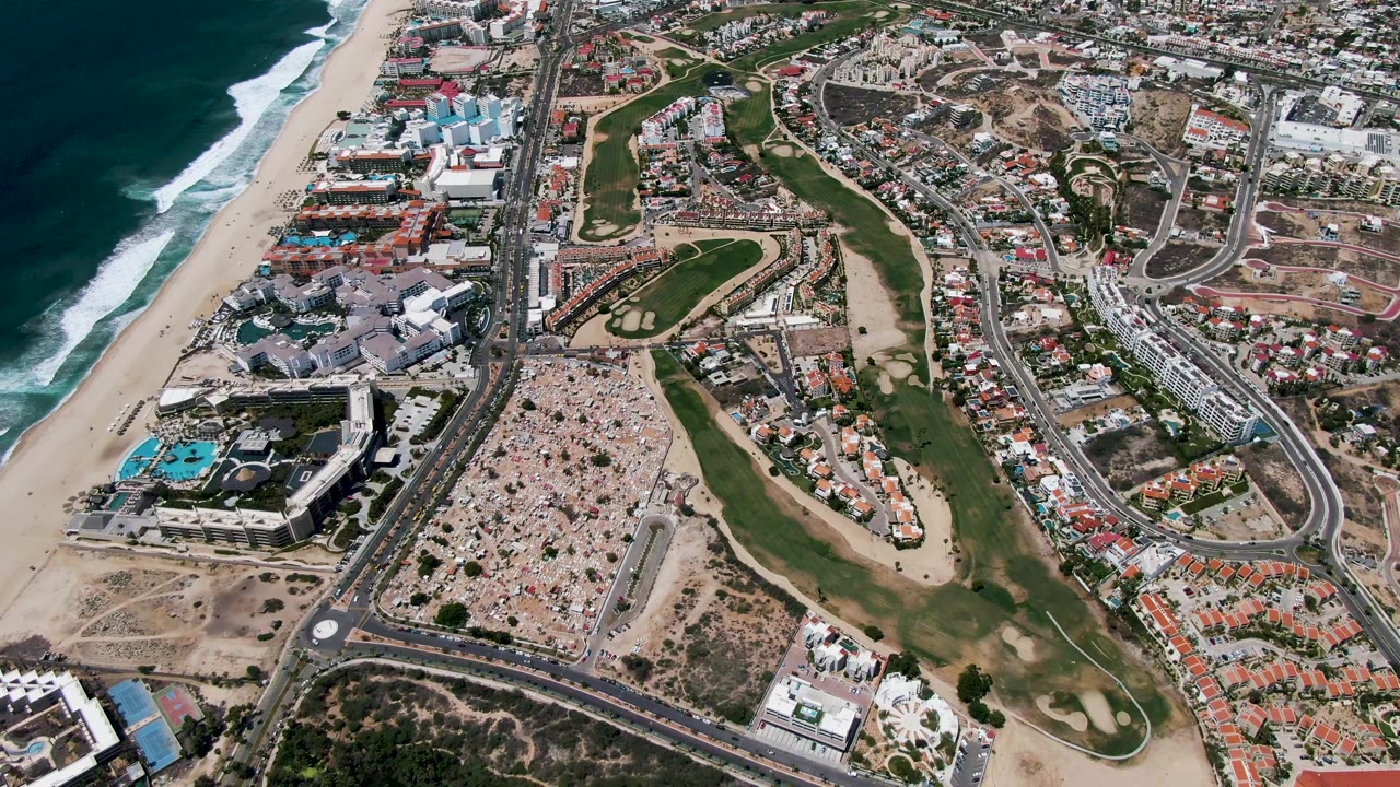 Opening aerial frame over San Jose Del Cabo, Mexico, with surf on the left, a sandy beach, green golf fairways, curved roads, pools, and no visible people.