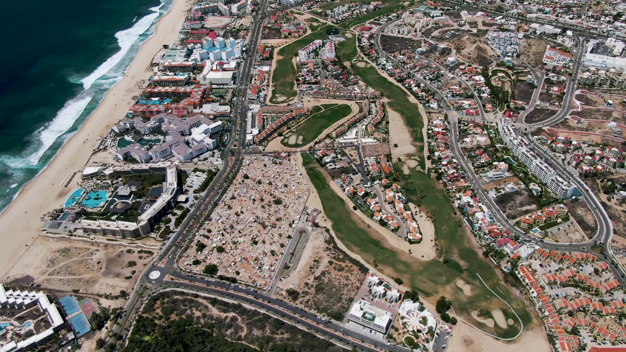 Second aerial frame of San Jose Del Cabo showing white surf bands along the beach, golf greens through the center, roadways, resort pools, and no visible people.