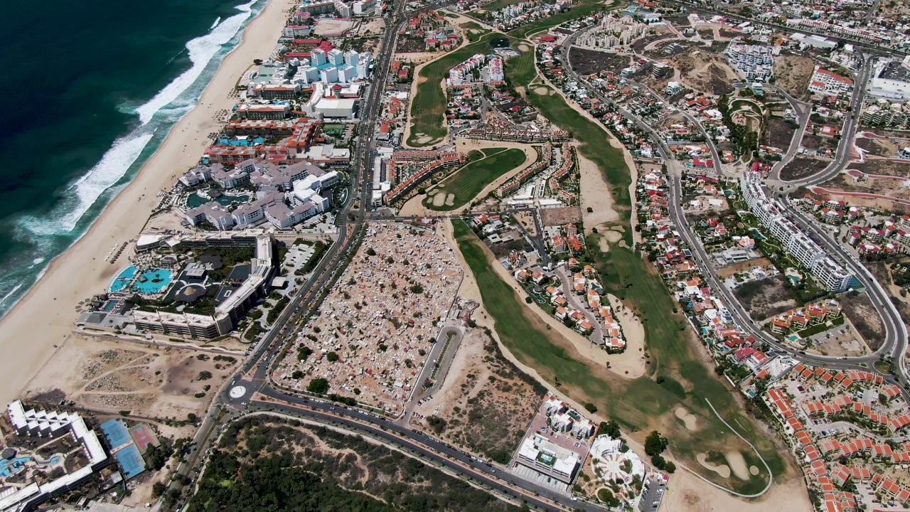 Third frame over San Jose Del Cabo with darker ocean water at left, breaking waves, the sandy shore, a central golf course, road grid, pools, and no visible people.