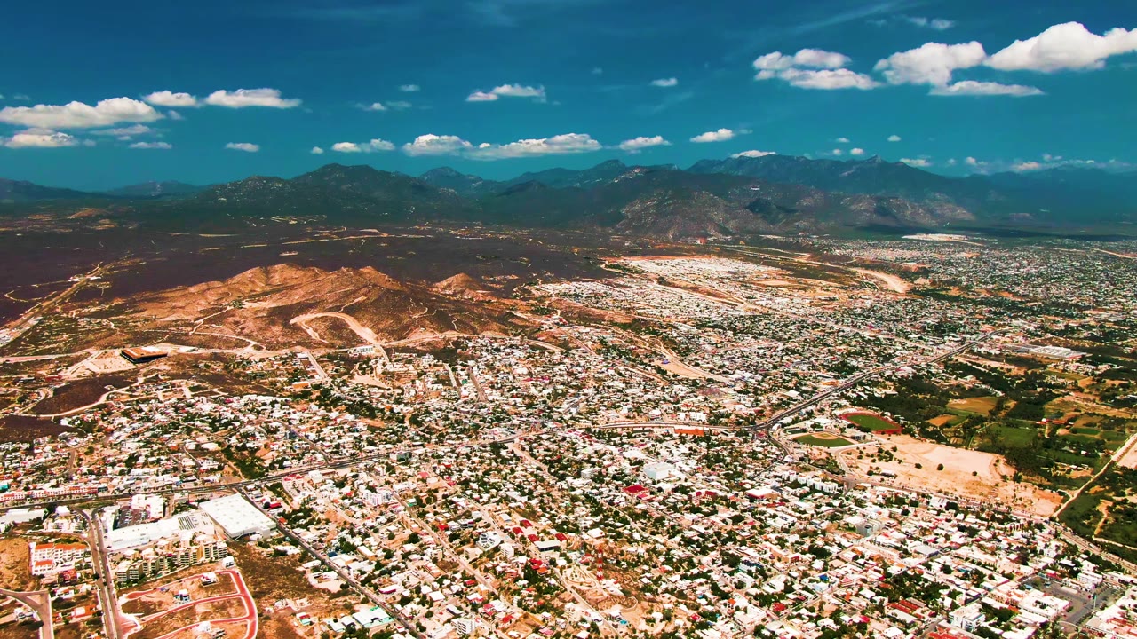 Wide aerial frame over San Jose del Cabo with low-rise buildings in the foreground, a tan hillside left of center, distant mountains, scattered white clouds, and no visible people.