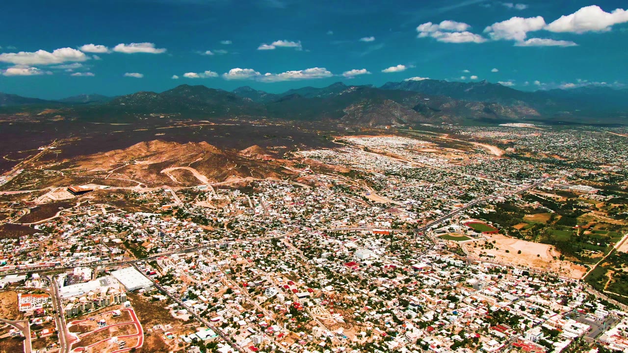 San Jose del Cabo appears from above with dense rooftops across the lower frame, pale open land to the right, rugged hills to the left, mountains in back, and no visible people.