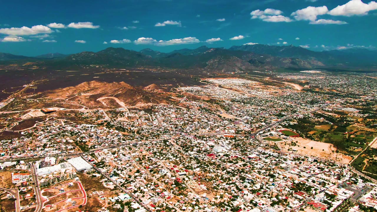 A high drone view over San Jose del Cabo shows a bright cityscape, a rounded tan hill near the center-left, road lines through the urban grid, distant mountains, and no visible people.