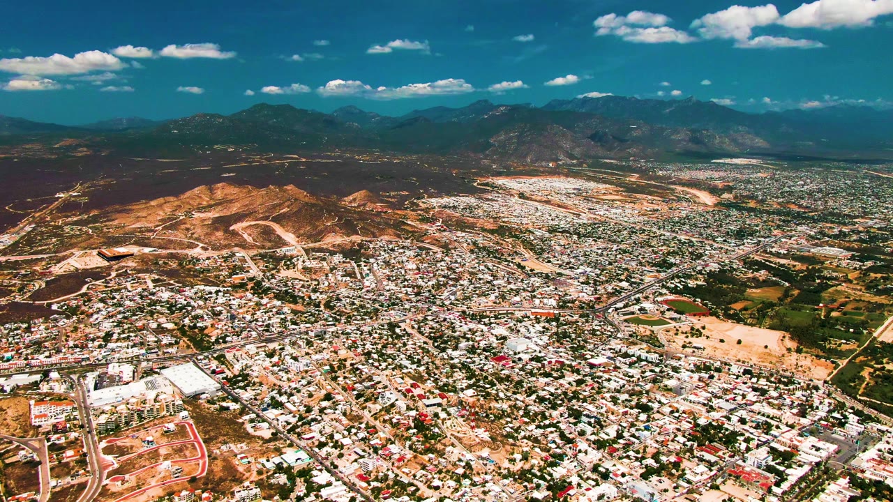 Aerial panorama of city blocks, brown hillside, green patches, mountains, and sky.