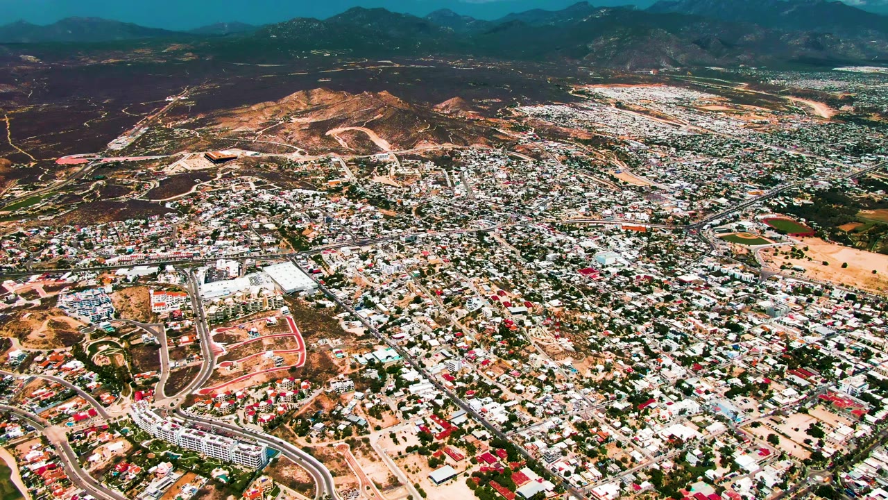 Opening aerial frame over San Jose Del Cabo, Mexico, showing dense low-rise development, curved roads, dry hills, and a dark mountain horizon with no visible people.