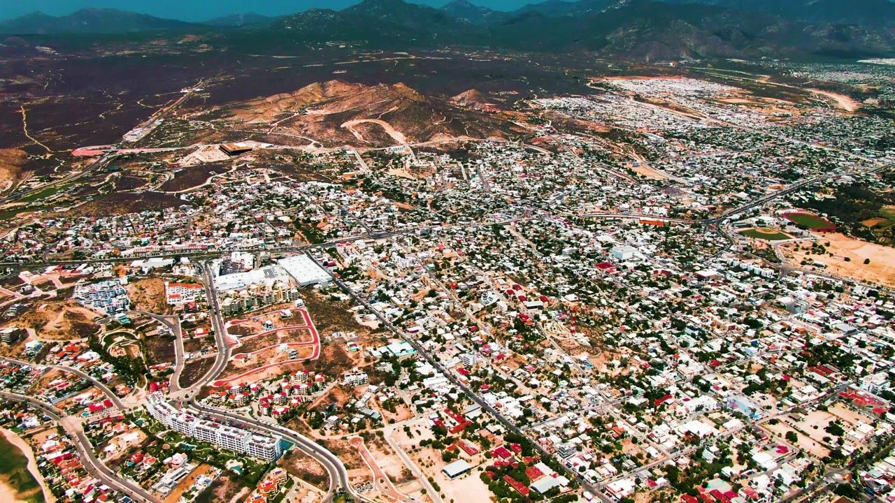 Second aerial frame of San Jose Del Cabo with a broad curved roadway in the lower area, dense buildings across the center, dry hills behind, and no visible people.