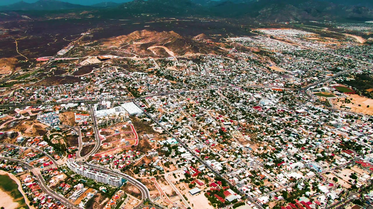 Third frame from the San Jose Del Cabo drone view, showing compact urban development, road curves near the lower frame, brown hillside terrain, and no visible people.