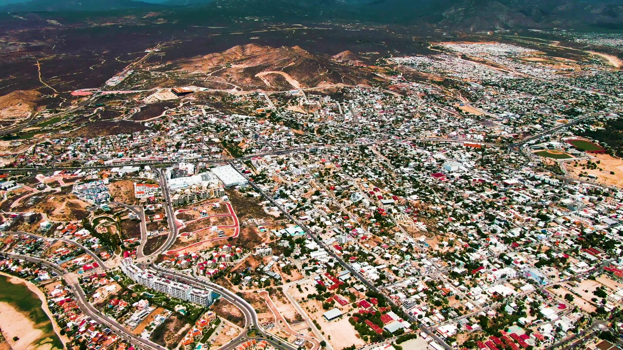 Aerial cityscape with dense rooftops, highway ramps, dry open land, and mountains.