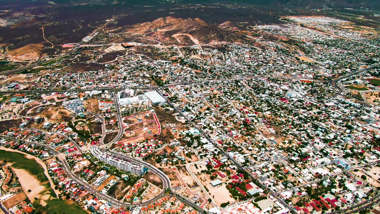 Aerial view over San Jose del Cabo with dense residential blocks, a curved road at lower left, green open areas, tan construction ground, distant hills, and no visible people.