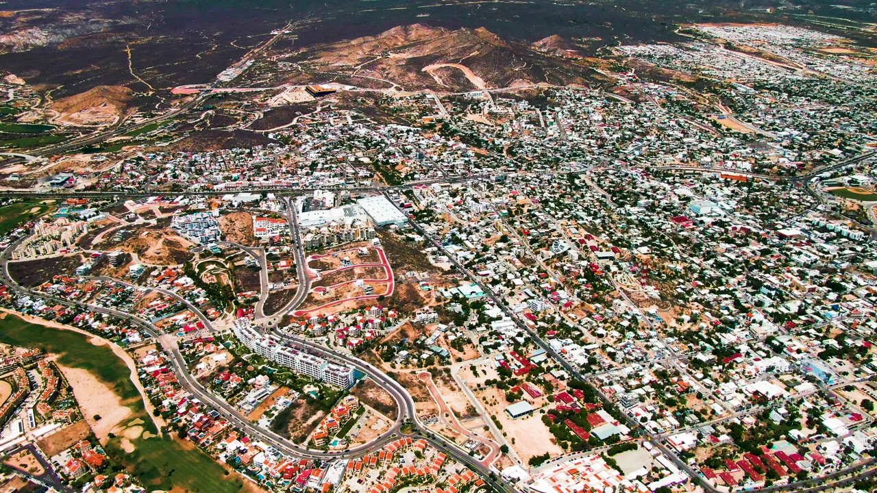 Aerial frame over San Jose del Cabo with residential streets filling the center, red and white rooftops, green terrain at the bottom edge, dry hills beyond town, and no visible people.