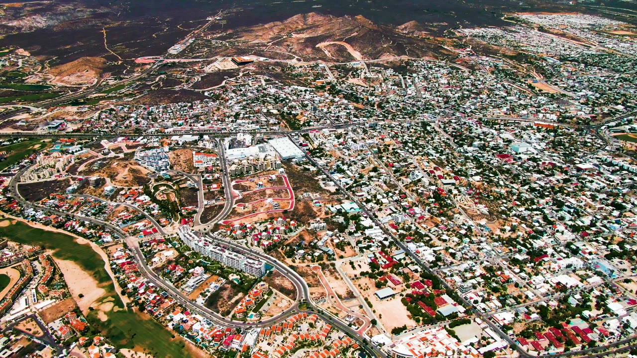 Wide aerial of suburban houses, curving roadway, and open hillside land.