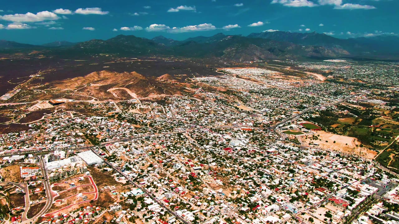 Wide aerial view over San Jose del Cabo with dense low-rise buildings, roads, tan hills, distant mountains, scattered clouds, and no visible people.