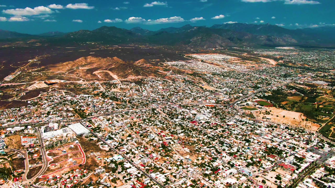 San Jose del Cabo appears from above with packed residential blocks, curving roads near the foreground, rust-colored hills, a blue mountain range, and no visible people.