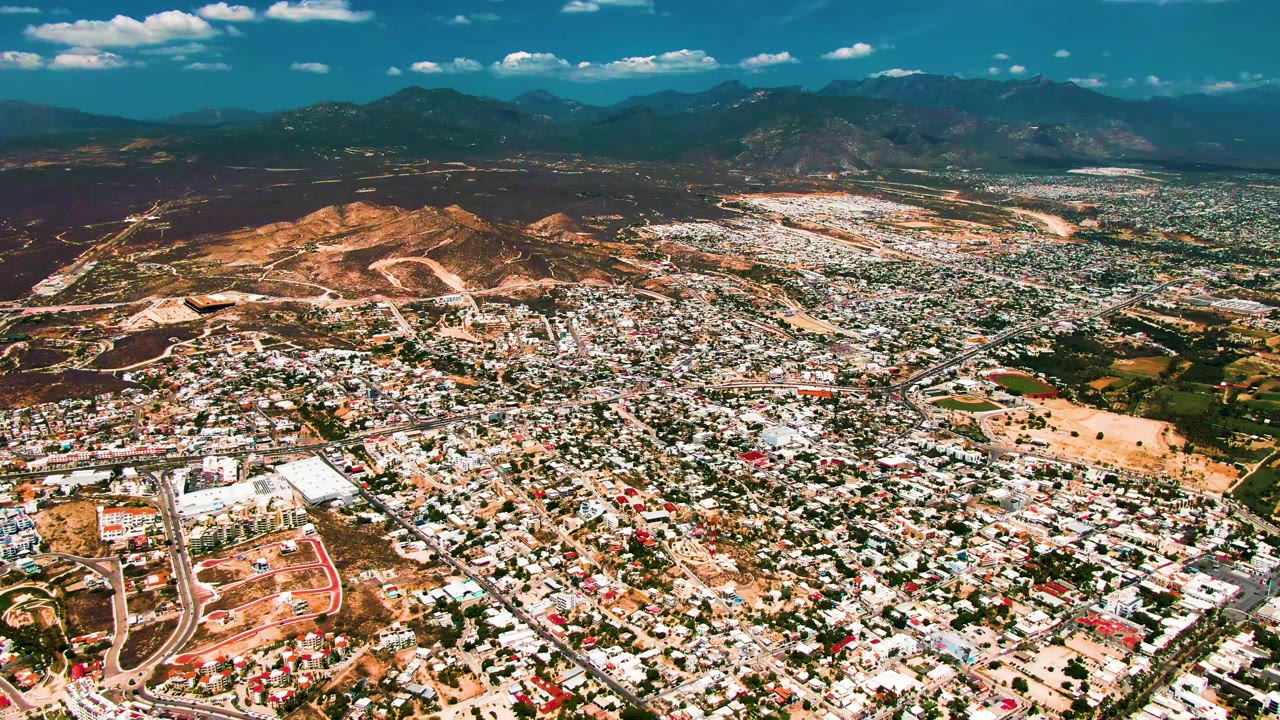 A wide San Jose del Cabo aerial frame shows rooftops and road lines across the foreground, dry open land and hills in the midground, mountains in the distance, and no visible people.