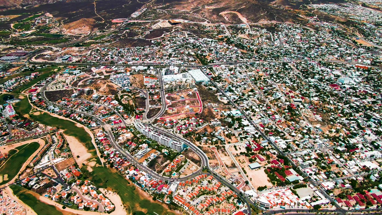 Aerial view over San Jose Del Cabo showing green golf fairways, sand traps, roads, compact housing, dry hills, and no visible people at the start of the clip.