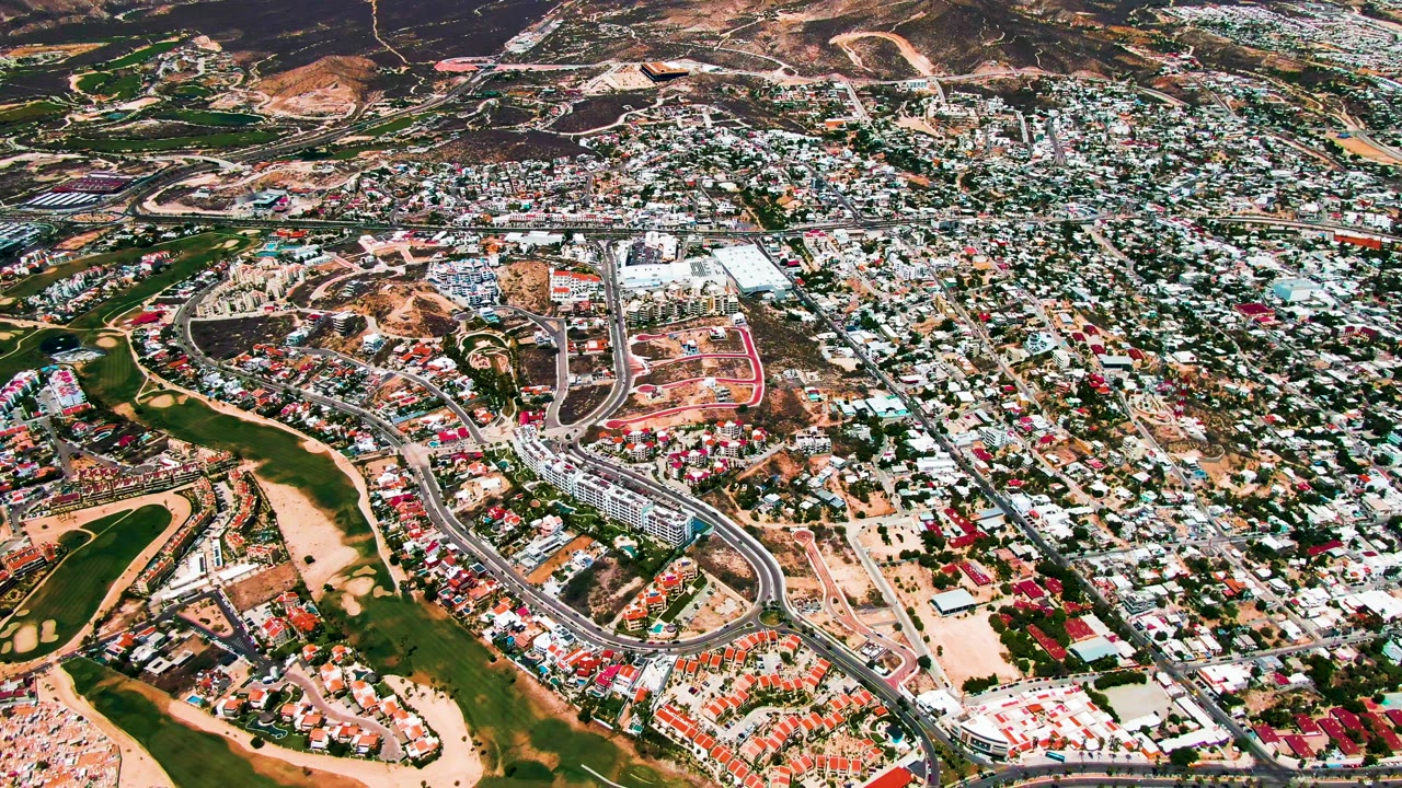 Drone aerial over San Jose Del Cabo with curving roads around the golf course, clusters of light rooftops, dry terrain beyond the city, and no visible people.