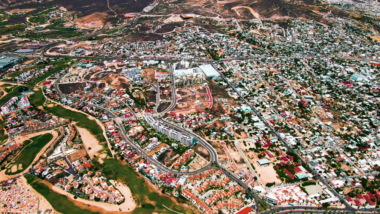 Overhead view of San Jose Del Cabo with the golf course crossing the lower frame, dense housing through the center, hillside terrain in the distance, and no visible people.