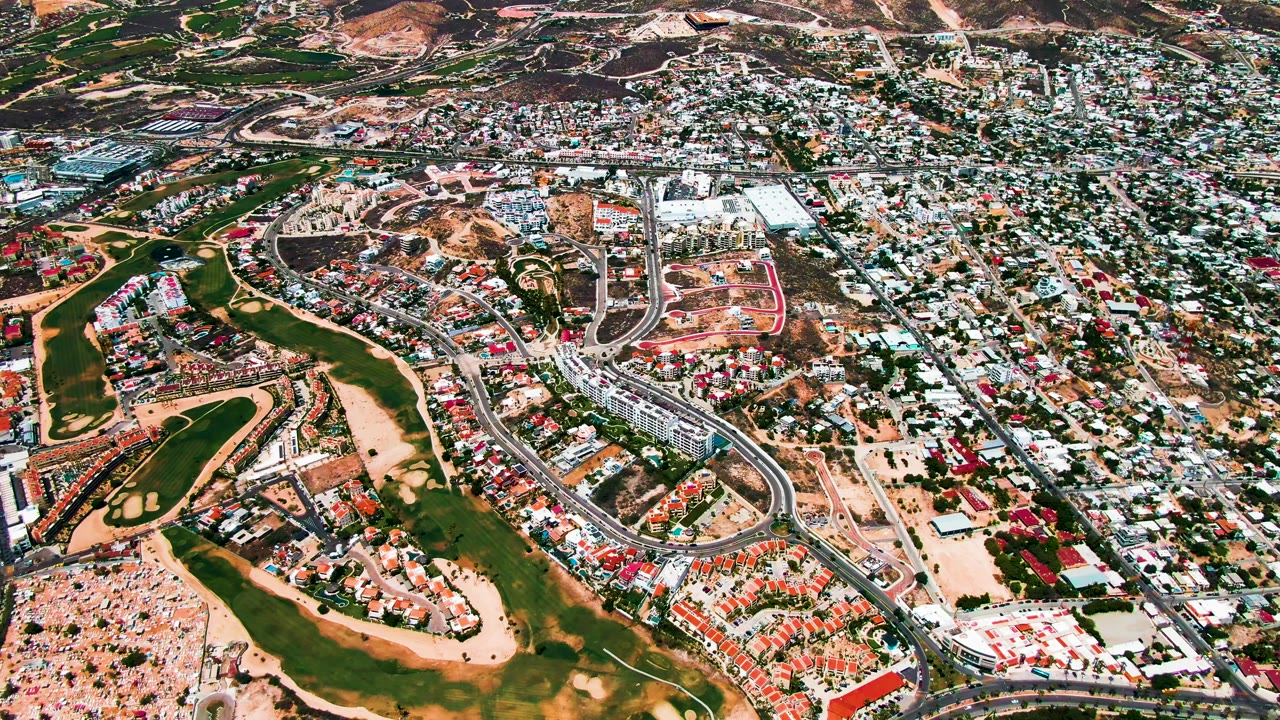 High aerial view over San Jose del Cabo showing green golf fairways, sandy rough, curving roads, dense houses, background hills, and no visible people.