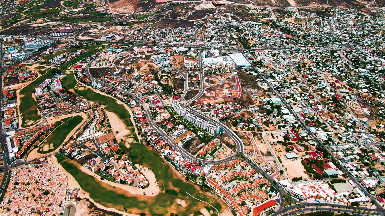 Aerial frame in San Jose del Cabo with fairways and tan ground in the lower left, a road corridor through the middle, compact housing to the right, and no visible people.