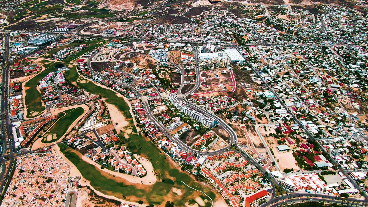 San Jose del Cabo aerial view with green course features at left, dense residential blocks at right, pale construction areas near the center, distant hills, and no visible people.