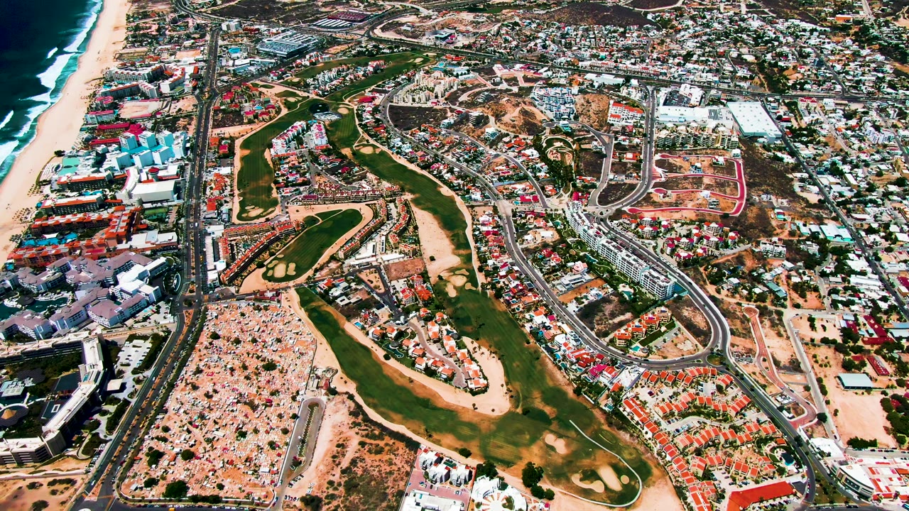 Opening aerial frame over San Jose Del Cabo with dark ocean and white surf at left, a sandy beach, green golf fairways, curved roads, parking areas, and dense urban development, with no visible people.