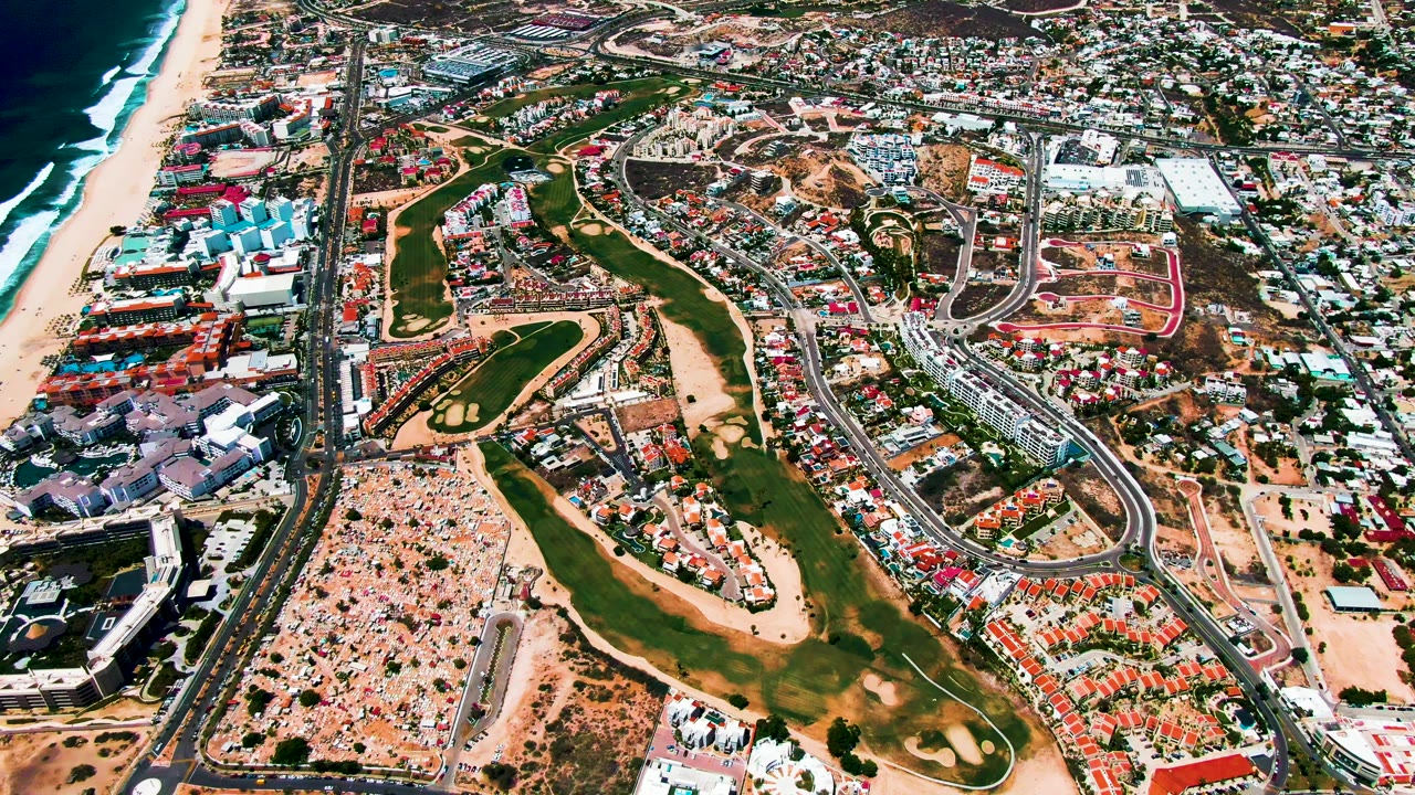 San Jose Del Cabo aerial view one second into the clip, with the beach and ocean still along the left edge, green fairways crossing the center, and roads and low-rise development filling the right side, with no visible people.