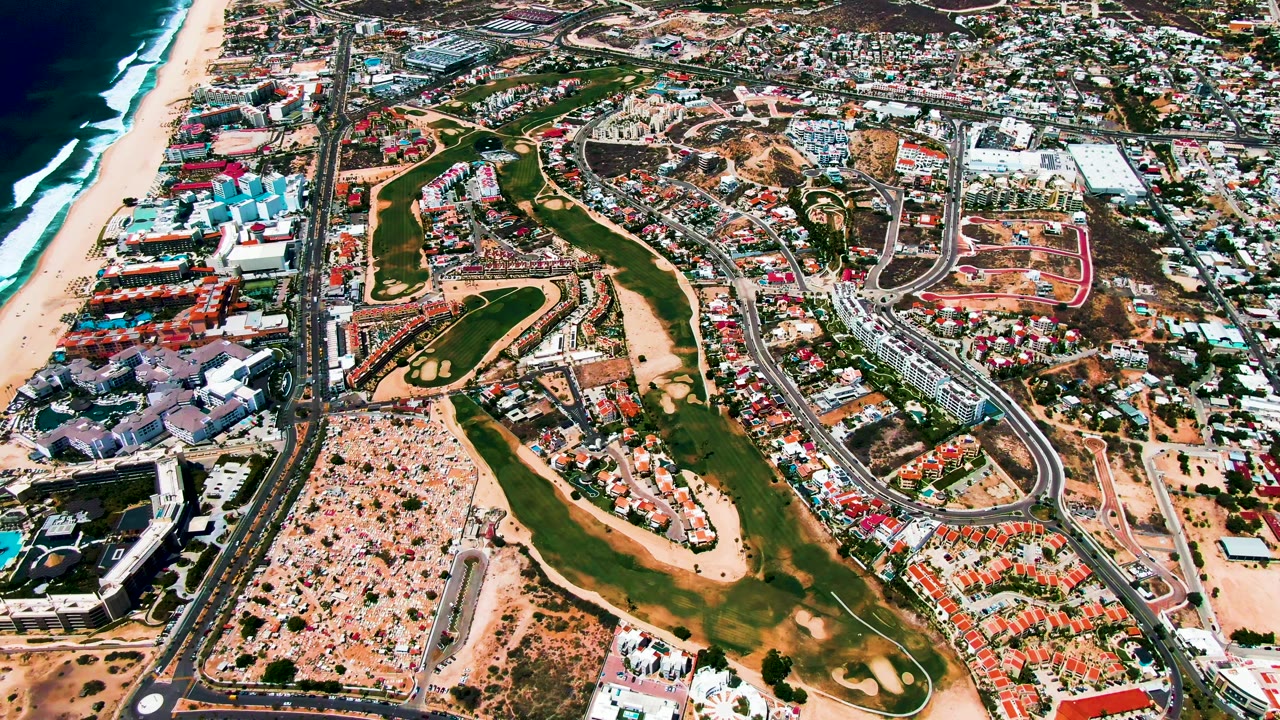 Aerial frame of San Jose Del Cabo showing ocean surf, a long sandy beach, resort-style buildings near the shoreline, green golf holes, road lanes, and compact town blocks, with no visible people.