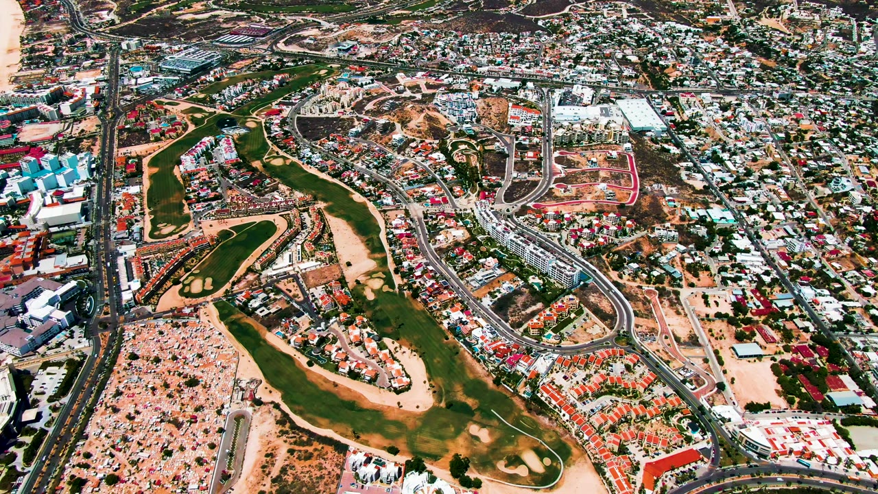 High aerial view over San Jose del Cabo with surf and beach at the right edge, a highway through the center, green fairway-like areas, residential blocks, and no visible people.