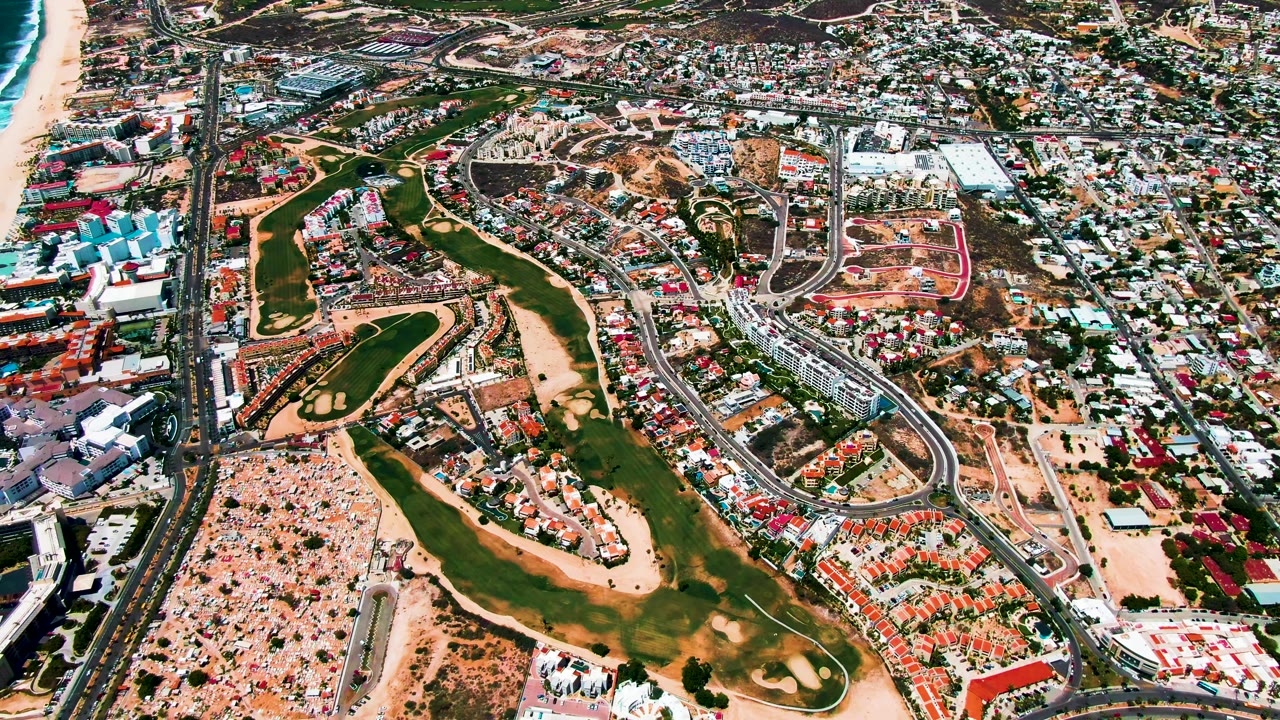 San Jose del Cabo aerial view with ocean surf at frame right, a long highway corridor, compact residential and commercial-looking buildings, green grounds, and no visible people.